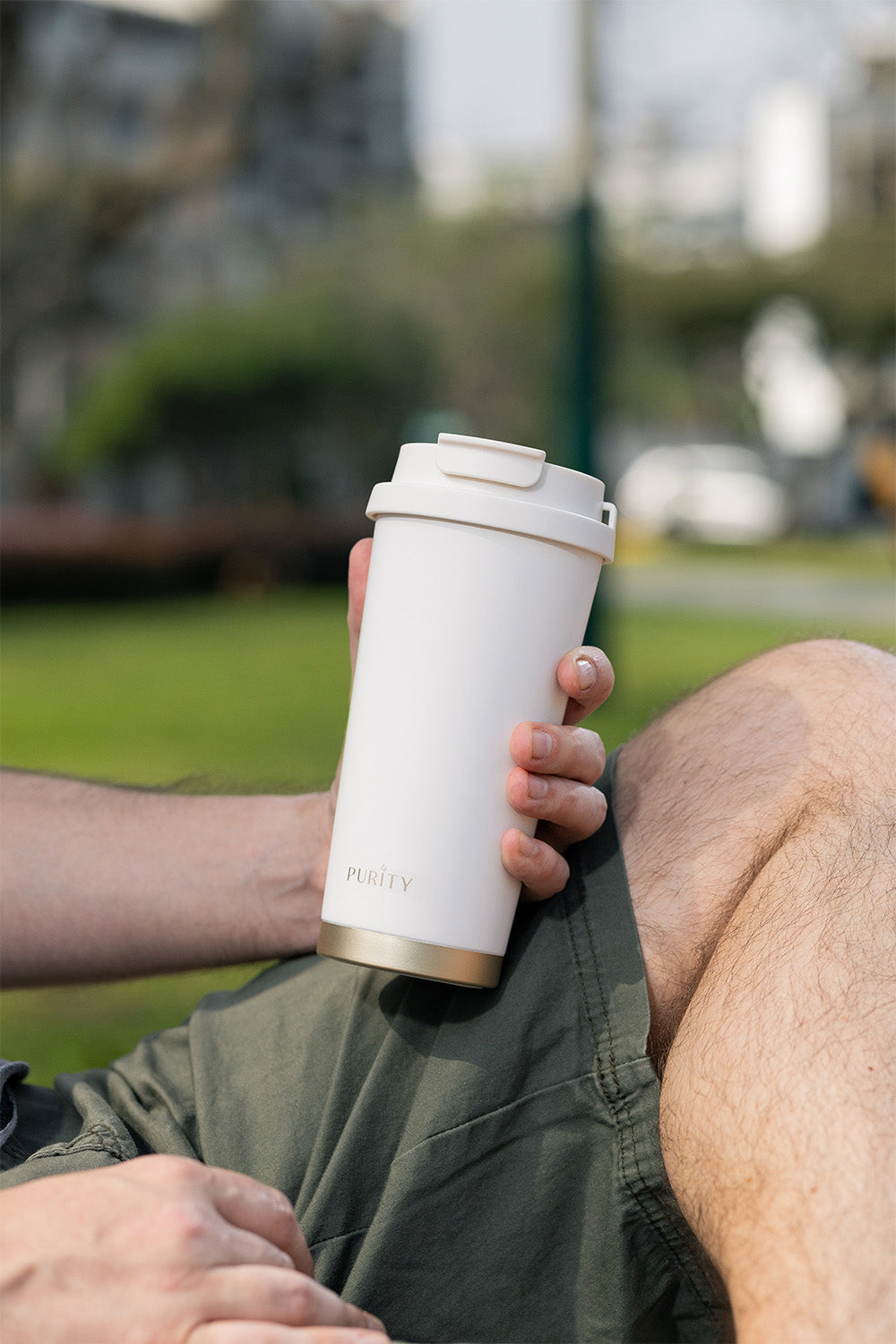 Una mano sostiene una taza de café Deluxe color blanco. En el fondo se ve un parque.