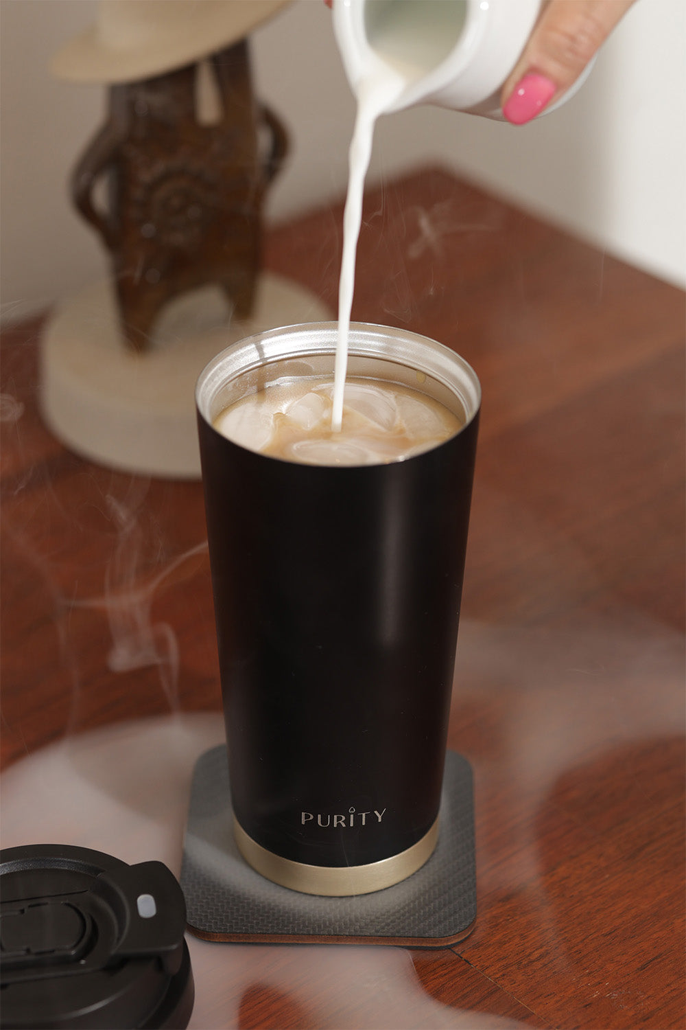 Taza de café Deluxe color negro destapado sobre un escritorio. Una mano vierte leche dentro de la taza.