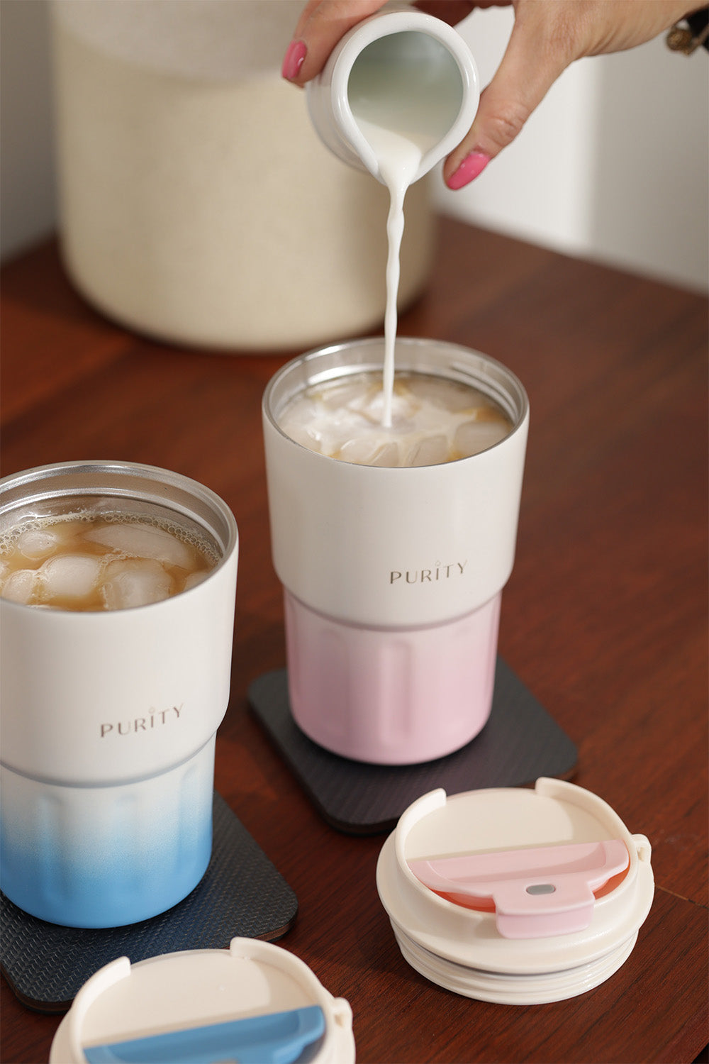 Una mano sirve leche en una taza Joy color rosado. a su lado hay otra taza Joy color celeste. Ambas están destapadas y llevan café helado en su interior. 